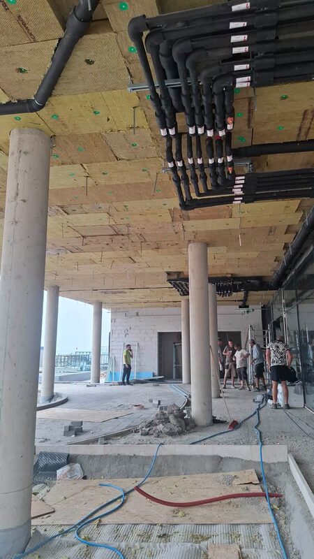Szigetelt gépészeti csőhálózat a födém alatt, betonoszlopos térben, a háttérben épülő medencetér